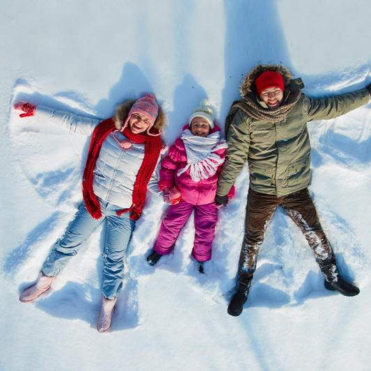 white mother and father lying down in snow with child between them, all making snow angels in snow and smiling www.shinethrivegrow.com