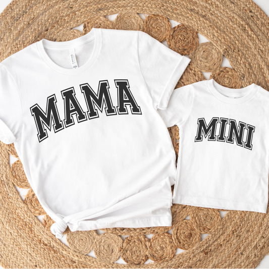 Photo of matching white short-sleeve t-shirts laying flat on rattan rug. Adult Mother's t-shirt has word "mama" on it. Letters are written in camo print. Child's t-shirt has word "mini" on it.  Letters are written in Capital letters in Varsity Letters font, black with white outline.